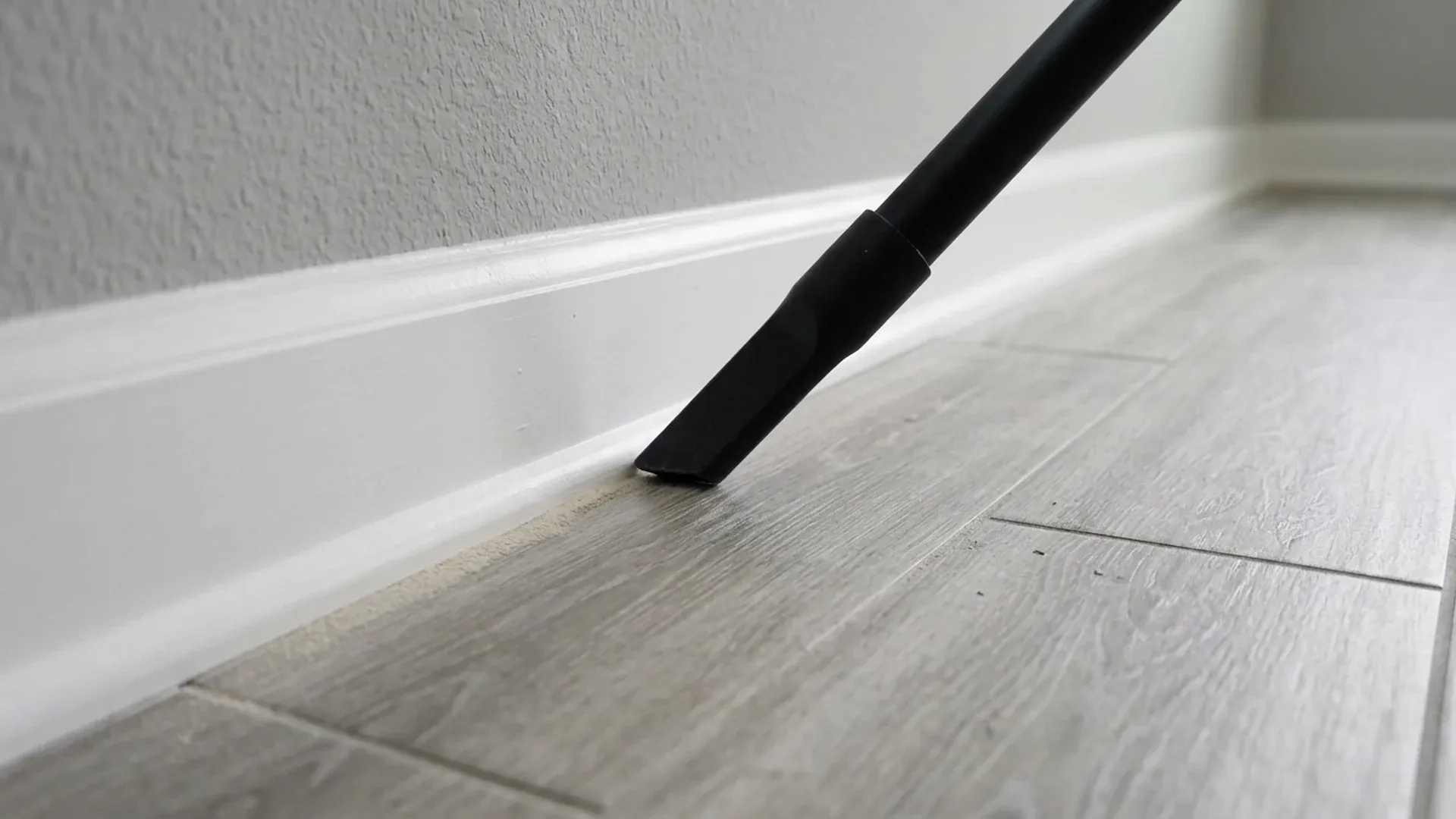 Vacuum crevice nozzle removing fine dust along a white baseboard beside a tile floor.